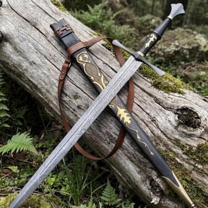 Puede incluir: Una espada de acero con hoja estampada y empuñadura negra con detalles dorados. La espada se acompaña de una vaina negra decorada con motivos de hojas doradas y una correa de cuero marrón, apoyada sobre un tronco de madera cubierto de musgo.