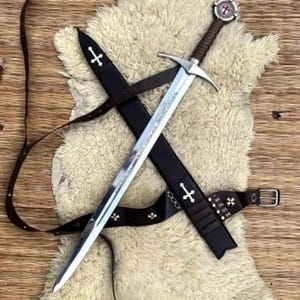 Pode incluir: Uma espada longa de aço com punho revestido em couro castanho e pomo circular com um emblema de cruz vermelha. A espada é acompanhada por uma bainha de couro preto e um cinto a condizer, ambos decorados com cruzes brancas e fivelas metálicas.