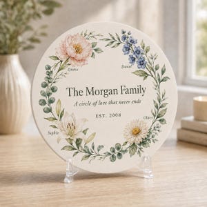 May include: A round, off-white ceramic plaque featuring a floral wreath illustration with pink, blue, and white flowers. The text reads The Morgan Family, A circle of love that never ends, EST. 2008, and the names Emma, Daniel, Oliver, and Sophie.