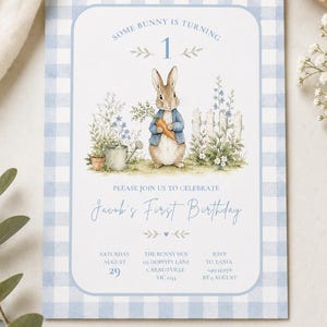 May include: A rectangular birthday invitation card with a light blue and white gingham border. The centre features a watercolour illustration of a rabbit in a blue jacket holding a carrot, surrounded by garden plants and a white fence. Text reads: SOME BUNNY IS TURNING 1, PLEASE JOIN US TO CELEBRATE Jacob's First Birthday, SATURDAY AUGUST 29, THE BUNNY DEN 123 HOPPITY LANE CARROTVILLE VIC 1234, RSVP TO TANIA 0412345678 BY 9 AUGUST.