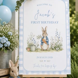 May include: A rectangular welcome sign for a first birthday featuring a blue gingham border. The text reads: Welcome to Jacob's First Birthday, 29 August 2026, and Thanks for hopping by. It displays a watercolour illustration of a rabbit holding a carrot.