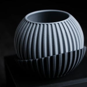Könnte beinhalten: Ein kugelförmiger Übertopf mit zweifarbigem Design, bestehend aus einem hellgrauen oberen Teil und einem dunkel anthrazitfarbenen unteren Teil. Die gesamte Oberfläche ist mit vertikalen, gleichmäßigen Rillen versehen, die eine gerippte Struktur erzeugen.