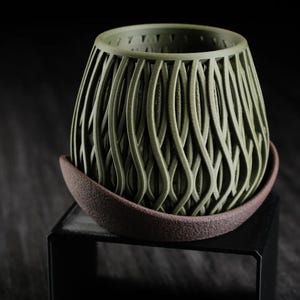 Peut inclure: Un pot de fleurs vert olive doté d'une structure en treillis tridimensionnelle complexe composée de brins verticaux, ondulés et interconnectés. Le pot repose dans une soucoupe brune texturée en forme de bol au bord incliné.
