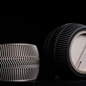 Op de afbeelding: Twee cilindrische objecten met een metallic afwerking en een uitgesproken geribbelde textuur met een herhalend golfpatroon. Het ene object staat rechtop, terwijl het andere gekanteld is om een gladde, ronde basis met een balk te tonen.