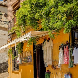 May include: A shop front with a bright yellow facade featuring a variety of hanging garments including white dresses, patterned skirts, and tote bags. A lush green vine grows over the entrance, which is shaded by a white fabric awning.