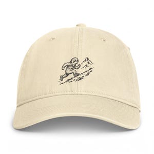 May include: A cream-colored baseball cap featuring a black line art illustration centered on the front panel. The graphic depicts a sloth wearing a backpack and hiking up a rocky mountain trail toward a mountain peak in the background.