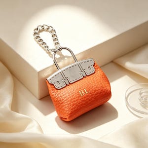 May include: A miniature orange textured handbag keychain featuring silver-toned metal hardware, a silver chain link attachment, and the letters BL embossed in gold on the front. The accessory has a structured shape with a flap and handle design.