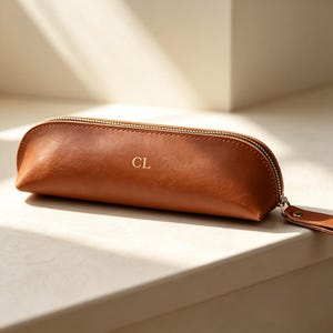May include: A tan, semi-cylindrical pencil case with a smooth finish and a gold-toned zipper closure. The letters CL are embossed in gold on the front center of the case. A matching tan leather-like pull tab is attached to the zipper slider.