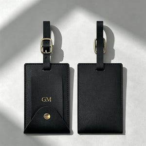 May include: Two rectangular black luggage tags with a textured finish. Each tag features a black strap with a gold-toned metal buckle. The left tag has a flap with a gold-toned snap button and the gold embossed initials GM on the front surface.