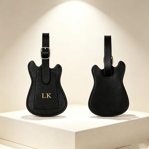 May include: Two black luggage tags shaped like electric guitars. One tag features the gold embossed letters LK on a textured surface and includes a silver-toned metal buckle on the strap. The other tag is plain black with a smooth finish.