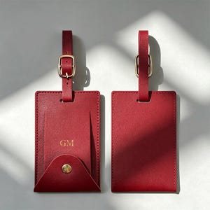 Pode incluir: Duas etiquetas de bagagem retangulares vermelhas com fivelas e alças douradas. Uma etiqueta possui uma aba presa por um botão de pressão dourado, com as letras GM gravadas em ouro. A outra é um retângulo vermelho liso com uma alça a condizer.