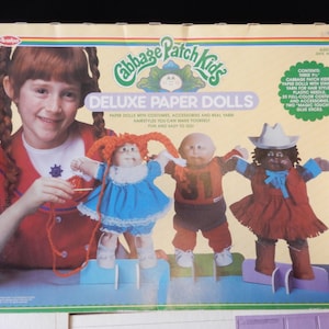 May include: A box of Cabbage Patch Kids Deluxe Paper Dolls. The box features a girl holding a doll and three dolls on stands. The dolls are wearing different outfits and have different hairstyles. The box includes a variety of accessories, including yarn, costumes, and a plastic needle.