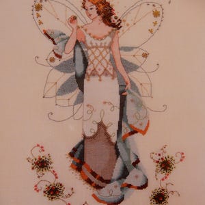 May include: Cross-stitch design of a fairy with auburn hair, wearing a white dress with gold accents and blue wings. The fairy is holding a small object. Floral embellishments surround the fairy. The background is a light beige color.
