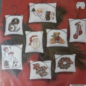May include: A Janlynn Christmas cross stitch kit featuring eight Holiday Dreams ornaments. Each ornament is a small, white pillow with a cross-stitched design, including a cat, angel, Santa, teddy bear, stocking, and wreath. Finished size: 3" x 4".