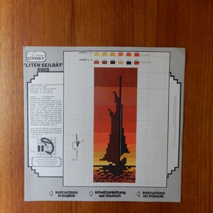 May include: A cross-stitch pattern for a sailboat, featuring a sunset color scheme with shades of orange, red, and brown. The design includes the text "Liten Seilbåt 6605" and instructions in English, German, and French.