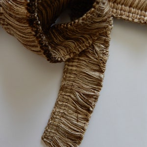 May include: Close-up of a decorative trim with a woven base and long, flowing fringe. The trim is a mix of light and dark brown threads, creating a textured appearance. The fringe adds a soft, tactile element to the design.
