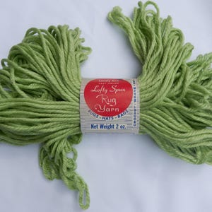 May include: A skein of light green "Lofty Spun Rug Yarn" with a red and white label. The label reads "Rugs, Hats, Bags" and "Net Weight 57 grams". The yarn is tightly wound and ready for crafting projects.