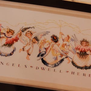 May include: Framed cross-stitch artwork featuring a group of angels with wings, holding flowers and ribbons. The text "ANGELS DWELL HERE" is stitched below the angels. The frame is light-colored wood.
