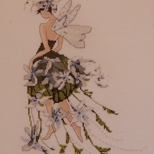 May include: Cross-stitch artwork of a fairy with a floral dress and wings. The fairy has a tan skin tone, dark hair, and is wearing a black top. The dress is green and blue, with white accents. The wings are white and blue.