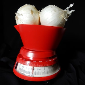 May include: Two white onions rest in a red bowl atop a vintage red kitchen scale. The scale's face displays measurements in grams and kilograms, with a maximum capacity of 2 kg. The background is black.