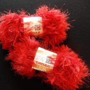 May include: Two balls of vibrant red yarn with a fluffy, textured appearance. Each ball has a label with the text "CIRCULO FRISE" and a QR code. The yarn is displayed against a black background, highlighting its color and texture.