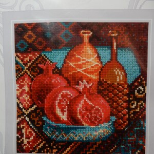 May include: Cross stitch kit featuring a still life of pomegranates and bottles on a patterned rug. The kit includes all the materials needed to complete the project. Stitch Kit.