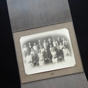 May include: A vintage black and white photograph of a group of people, likely from the early 20th century, is displayed within a textured gray photo album. The album is open, revealing the photograph and its decorative border.