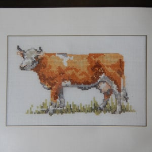 May include: Framed cross-stitch of a brown and white cow standing in a field of green grass. The cow is detailed with shades of brown, white, and gray. The artwork is framed in a black frame with a white mat.