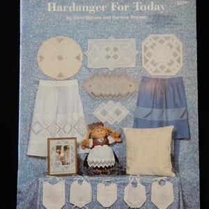 May include: A book cover titled "Hardanger For Today" featuring various white embroidered items, including a skirt, placemats, and a pillow. A doll in traditional clothing and framed embroidery are also displayed. The background is a light blue floral pattern.