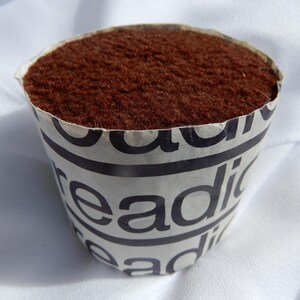 May include: A small, round, brown object with a textured surface, set inside a paper cup. The cup is white with black text that reads "readi". The object is likely a pincushion, used for holding sewing needles.