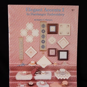 May include: Book cover for "Elegant Accents I in Hardanger Embroidery" by Rosalyn K. Wetnamo. The cover features a pink background with white polka dots and various examples of Hardanger embroidery projects, including bookmarks, small squares, and a napkin.