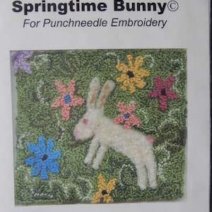 May include: Punchneedle embroidery pattern featuring a white bunny surrounded by colorful flowers on a green background. The pattern is titled "Springtime Bunny" and is for punchneedle embroidery. The finished size is 3 1/2 inches by 3 inches.