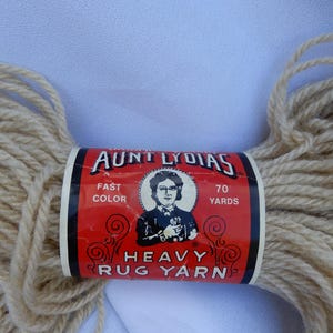 May include: Aunt Lydia's Heavy Rug Yarn, a beige yarn skein with a red label. The label features a portrait of a woman and the text "Fast Color" and "70 Yards". The yarn is designed for rug making.