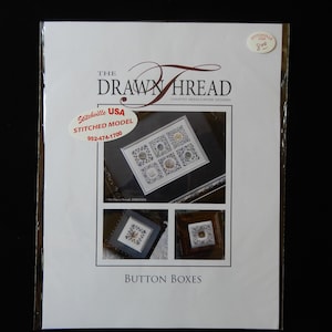 May include: A packaged cross-stitch pattern from "The Drawn Thread" featuring "Button Boxes." The package includes a stitched model and the Stitchville USA contact information. The design is displayed in a clear plastic sleeve.