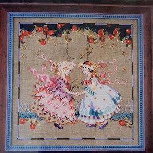 May include: Cross-stitch artwork featuring two girls in dresses, holding hands. The girls are under a vine with red apples. The artwork is framed in a dark brown wooden frame with a blue border.