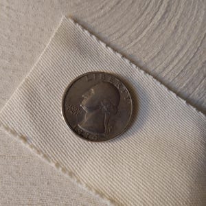 May include: A silver quarter coin from 1974 rests on a piece of off-white fabric, with a roll of paper in the background. The coin features a profile of George Washington and the words "In God We Trust" and "Liberty".