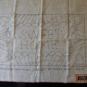 May include: A tan burlap fabric piece featuring a black line drawing of a rectangular border with hearts in the corners and the text SWEET HOME in the center. Below the design, the text reads PINE ISLAND PRIMITIVES Sally Kallin 18x31.