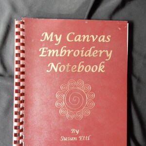 May include: A red spiral bound notebook with the title "My Canvas Embroidery Notebook" in gold lettering. The cover features a gold design of a circle with swirls. The author's name, "Susan Ettl", is printed in gold lettering below the design.