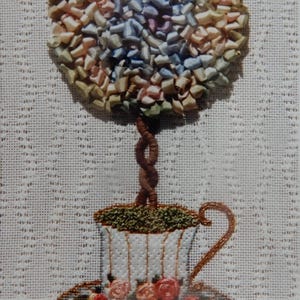 May include: Embroidered artwork featuring a topiary tree in a teacup. The tree's foliage is made of colorful ribbon, and the teacup is decorated with roses. The artwork is on a white, textured background.