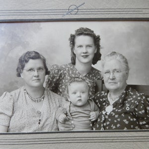 May include: Black and white photo of four people, three women and a young child. The women are wearing patterned dresses and the child is wearing a striped shirt. The photo is framed in a simple black frame.