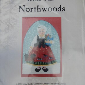 May include: A cross-stitch pattern featuring "Little Ms. Northwoods." The design shows a woman in a red dress holding gifts and a basket. The background is a light blue with a red arch. The text "Little Ms. Northwoods" is at the top.