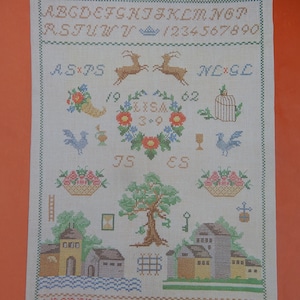 May include: Cross-stitch sampler with a cream background, featuring an alphabet, numbers, and various embroidered images. Includes deer, floral wreaths, birds, houses, and baskets. The dimensions are 11 3/4 x 17 inches.
