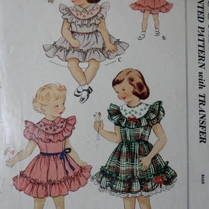 May include: Vintage sewing pattern featuring illustrations of little girls in dresses. The dresses have ruffled sleeves and skirts, with details like floral embroidery and plaid patterns. The pattern includes the text "PRINTED PATTERN with TRANSFER" in blue.