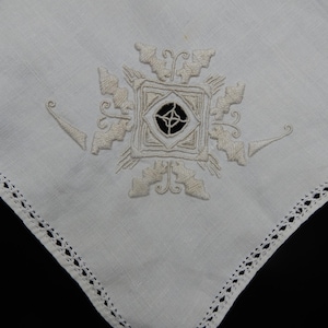 May include: White linen handkerchief with an embroidered central design. The design features a square with a black center and ornate, cream-colored floral and geometric details. The edges are finished with a delicate, openwork trim.