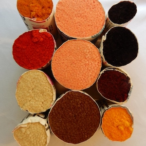 May include: A collection of colorful yarn skeins in various shades, including orange, red, peach, brown, and cream. The yarn is tightly wound and presented in cylindrical paper packaging, ideal for knitting or crochet projects.