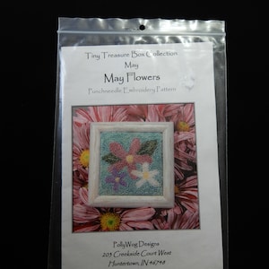 May include: A punch needle embroidery pattern for May Flowers, part of the Tiny Treasure Box Collection. The pattern features two flowers in a white frame against a background of pink flowers.