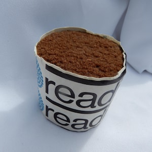 May include: A small, round object with a brown, textured top, set in a paper cup. The cup is white with black text that reads "read" and blue droplet designs. The object is on a white surface.