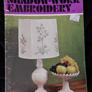 May include: A vintage book titled "Shadow-Work Embroidery" with a white lampshade with a floral design, a white bowl with grapes and apples, and a green box on a wooden surface.