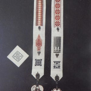 May include: A white embroidered textile with intricate red and black geometric patterns. The design includes a long, narrow scarf-like piece with decorative ends, a heart-shaped pendant, and a square piece.