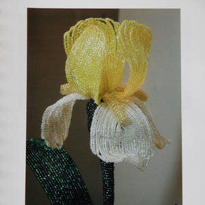 May include: A beaded iris flower sculpture with yellow upper petals and white lower petals, featuring a dark green beaded stem and leaf. The text "French Beaded Iris" appears at the top and "By Donna DeAngelis Dickt" at the bottom.